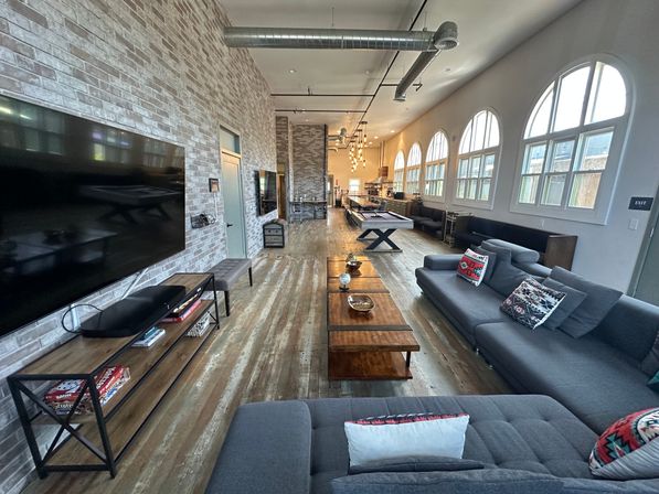 Spacious industrial-style lounge and game room with exposed ductwork, brick accent wall, arched windows, wood floors, large sectional sofa, wall TV, and pool table