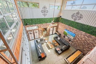 Sunlit industrial-style loft living room with high ceilings, brick accent wall, floor-to-ceiling glass garage doors, black leather sectional, wall-mounted TV, shuffleboard table and greenery trim.