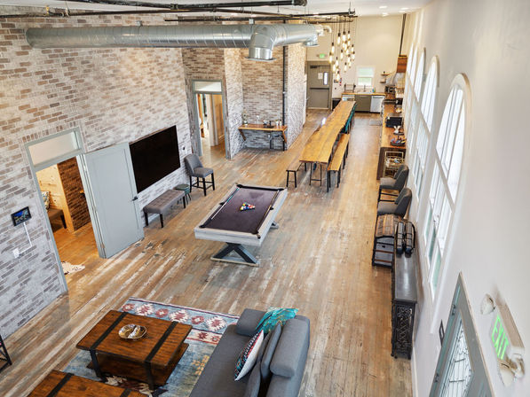 Loft-style communal lounge with pool table, long farmhouse table, exposed brick walls, industrial ductwork, arched windows, worn hardwood floors and cozy seating.