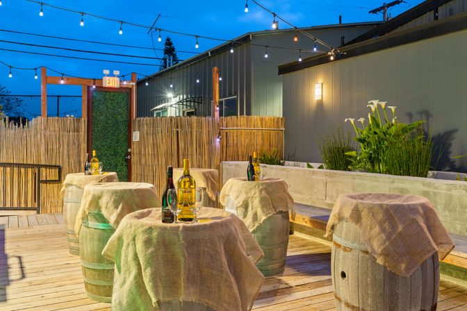 Outdoor wooden patio at dusk with wine-barrel tables draped in burlap, wine bottles and glasses on top, string lights overhead, bamboo fence and potted greenery.