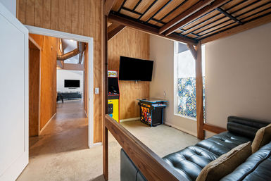 Cozy loft-style game room with wood-paneled walls, wall-mounted TV, retro arcade machines and cocktail cabinet, black leather sofa beneath a wooden loft bed, patterned window shade and concrete floor