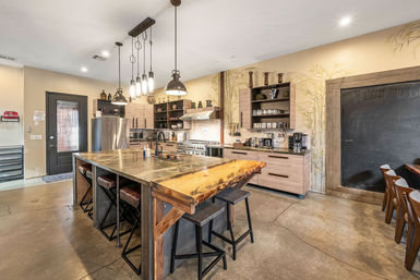 Modern rustic open-concept kitchen with large stone and live-edge wood island, bar stools, industrial pendant lights, stainless-steel appliances, light wood cabinets, chalkboard accent wall and polished concrete floors.