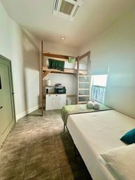 Cozy studio bedroom with wooden loft bunk over a small kitchenette, ladder access, full bed with rolled towels, tile floor and a sunlit window