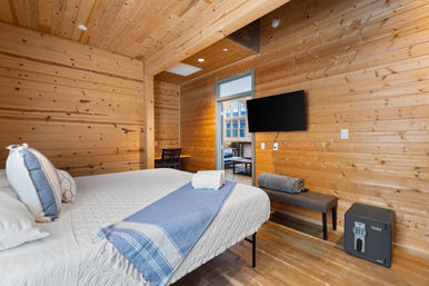 Cozy wood-paneled cabin bedroom with white linens and blue throw on bed, wall-mounted TV, bench and digital safe, desk and seating visible through doorway