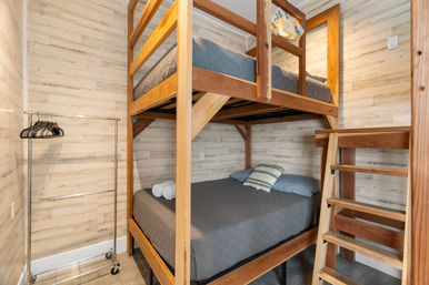 Cozy compact bedroom with a wooden loft-style bunk bed, ladder, gray bedding, striped accent pillow, rolled towels and a rolling garment rack against light shiplap walls.
