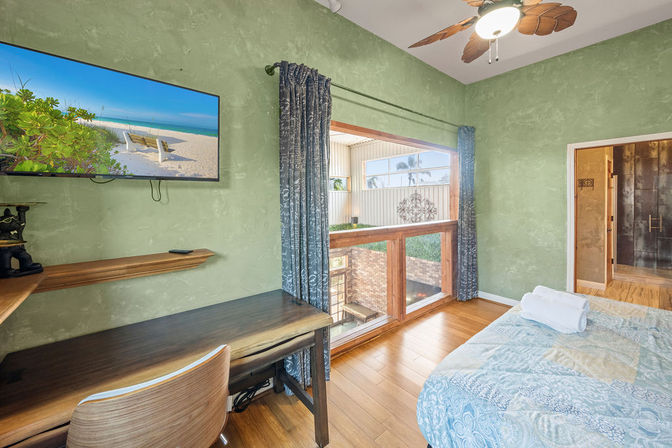 Coastal-themed bedroom with green textured walls, hardwood floors, wooden desk and chair, wall-mounted TV showing a beach, large wood-framed window opening to a sunlit patio/garden, blue patterned curtains, leaf-bladed ceiling fan and a bed with a folded towel.
