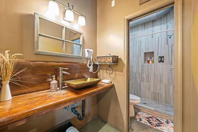 Cozy rustic bathroom with wooden live-edge vanity, bronze vessel sink and faucet, wall-mounted mirror and lights, hairdryer and toiletries, and adjacent tiled walk-in shower with built-in niche.