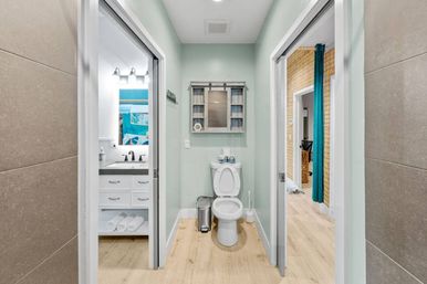 Narrow modern bathroom alcove with white toilet centered beneath a rustic wall cabinet, mint-green walls and light wood-look floor; left doorway shows a white vanity and mirror, right doorway reveals an adjacent room with a teal curtain.
