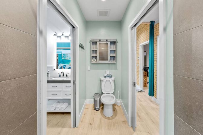 Narrow modern bathroom alcove with white toilet centered beneath a rustic wall cabinet, mint-green walls and light wood-look floor; left doorway shows a white vanity and mirror, right doorway reveals an adjacent room with a teal curtain.