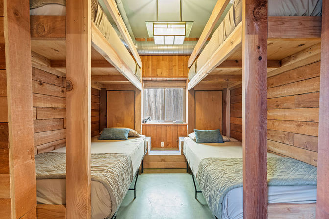 Cozy wood-paneled cabin bunk room with two sets of twin bunk beds, neutral quilts and pillows, a central window and warm natural light