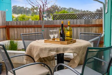 Cozy fenced backyard patio with round table draped in burlap, wooden tray holding red and white wine bottles and two wine glasses, surrounded by four metal mesh chairs.