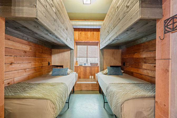 Cozy rustic cabin bunk room with twin beds facing each other, reclaimed wood-paneled walls and overhead loft bunks, small center window, green quilts and blue accent pillows.