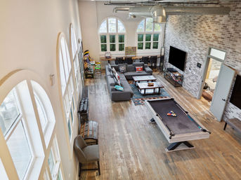Spacious industrial-style loft game room with large arched windows, exposed brick wall, hardwood floors, gray sectional sofa, coffee tables, desk area and a pool table