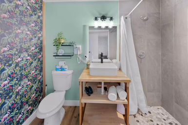 Small modern bathroom with flamingo tropical wallpaper, mint-green wall, wooden vanity with white vessel sink and mirror, and gray-tiled walk-in shower with white curtain.
