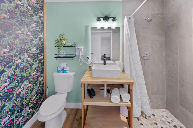 Small modern bathroom with flamingo tropical wallpaper, mint-green wall, wooden vanity with white vessel sink and mirror, and gray-tiled walk-in shower with white curtain.