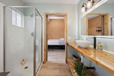 Bright modern-rustic bathroom with glass walk-in shower, wood floating countertop, white vessel sink, large mirror, potted plant and doorway to a wood-paneled bedroom
