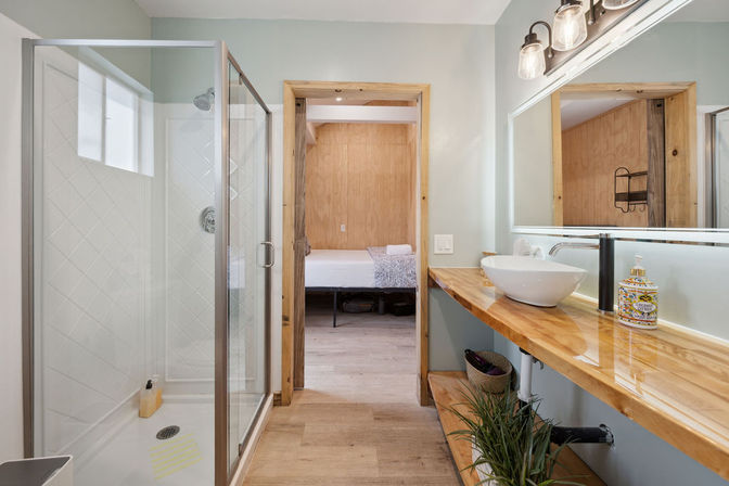 Bright modern-rustic bathroom with glass walk-in shower, wood floating countertop, white vessel sink, large mirror, potted plant and doorway to a wood-paneled bedroom