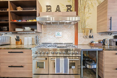 Stainless-steel professional range and hood centered in a bright modern kitchen with wood cabinets, open shelving of colorful dishes, subway tile backsplash, hanging utensils, and countertop appliances