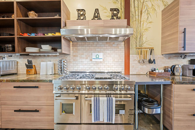 Stainless-steel professional range and hood centered in a bright modern kitchen with wood cabinets, open shelving of colorful dishes, subway tile backsplash, hanging utensils, and countertop appliances
