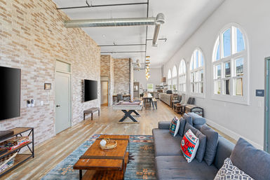 Bright industrial loft lounge with exposed brick wall, arched windows, hardwood floors, open kitchen, pool table and a cozy sofa with colorful pillows.