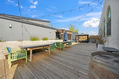 Inviting outdoor wooden deck patio with long concrete-top tables, lime green cushioned chairs, string lights overhead, umbrella and shaded lounge seating, rustic barrel planters and blue sky.