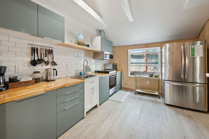 Bright contemporary kitchen interior with sage-green cabinets, butcher block countertops, white subway-tile backsplash, stainless steel French-door refrigerator and gas range, sink by a window and light wood plank flooring.
