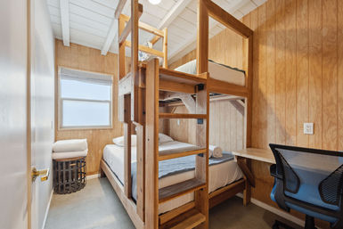 Cozy cabin-style wood-paneled bedroom with sturdy wooden bunk bed and ladder, crisp white linens and rolled towel, small frosted window, wicker basket with folded towels, and a compact desk with mesh office chair.