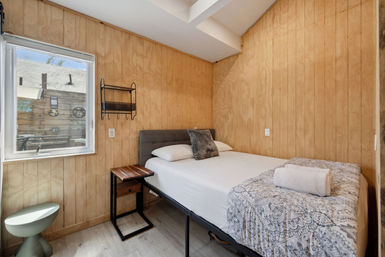 Cozy small wood-paneled bedroom with skylight and window, queen bed with gray headboard, patterned throw and rolled towel, bedside table, wall shelf and light wood floors.