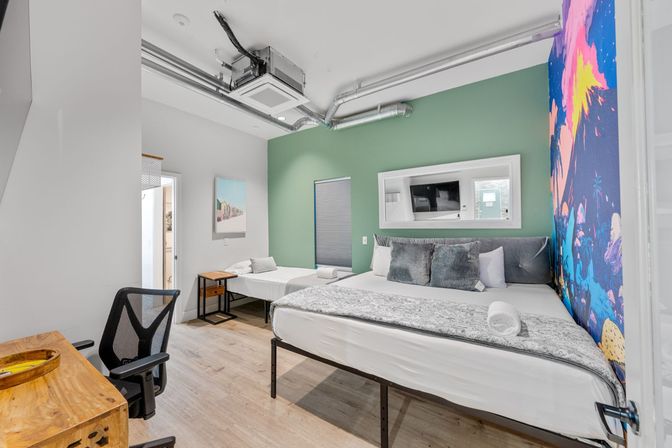 Modern urban guest room with king bed and single daybed, green accent wall, colorful tropical mural, exposed ductwork, desk, and wall-mounted TV.