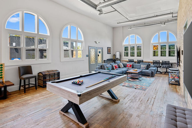 Bright loft-style game room and lounge with a modern pool table, gray sectional sofa with colorful pillows, arched windows, hardwood floors, and exposed ductwork