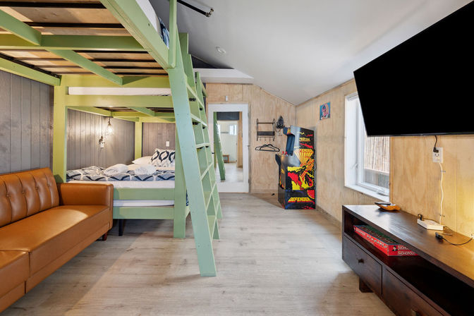 Bright loft-style bunk room with mint-green wooden bunk beds and ladder, brown leather sofa, retro arcade machine, wall-mounted TV, light wood floors and wood-paneled walls — cozy family-friendly rental space.