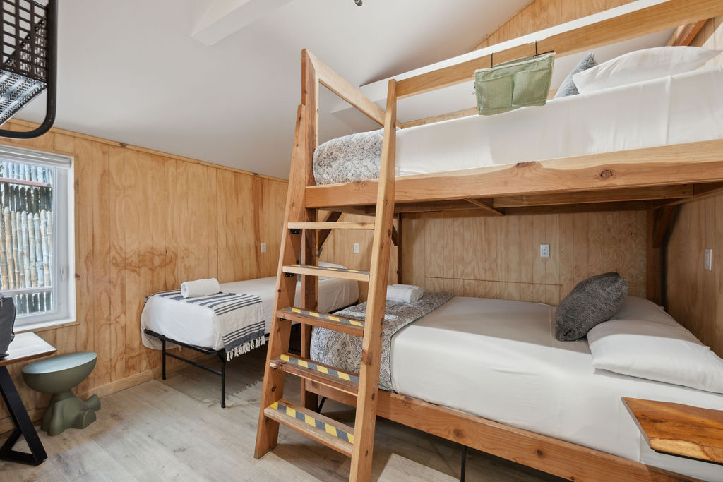Cozy rustic bunk room with light wood paneling, ladder-access wooden bunk beds, a single bed by the window, neutral linens and a small desk.