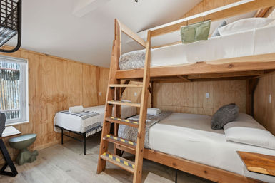 Cozy rustic bunk room with light wood paneling, ladder-access wooden bunk beds, a single bed by the window, neutral linens and a small desk.