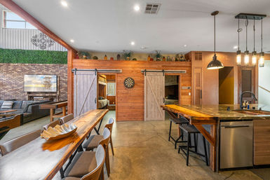 Modern rustic open-concept loft interior with live-edge wood dining table, kitchen island with pendant lights and bar stools, sliding barn doors to bedrooms, dartboard on wood-paneled wall, exposed brick accent and sectional seating.