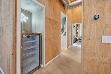 Wood-paneled hallway interior with concrete floor and exposed metal ductwork, featuring a glass-door beverage fridge under a neon surfboard sign and doorways leading to a coastal-themed living area.