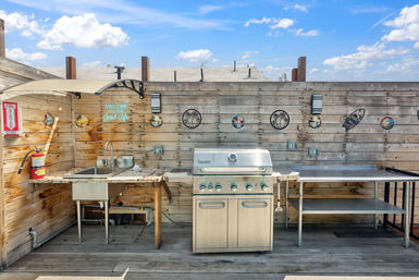 Sunny rooftop patio outdoor kitchen with stainless steel gas grill, sink and prep counters against a wood plank fence decorated with tropical wall art.