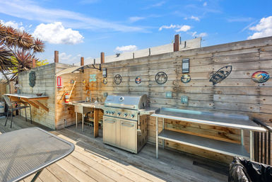 Rooftop outdoor kitchen with stainless-steel gas grill, prep tables and sink against a weathered wooden fence decorated with surf and palm artwork, tropical plants and a bright blue sky