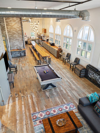Bright industrial farmhouse loft with a pool table on reclaimed wood floors, long communal dining table, modern kitchen cabinets and arched windows flooding the space with natural light.