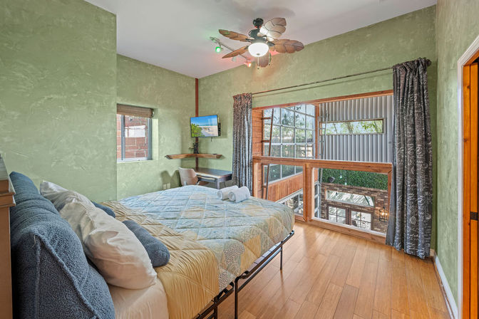 Cozy loft-style bedroom with green textured walls, hardwood floors, quilted bed with blue pillows, leaf-blade ceiling fan, small desk and wall-mounted TV, and large interior window railing overlooking a sunlit wood-and-metal atrium.