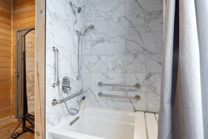 Accessible tub-shower with white marble-pattern tile, chrome overhead and handheld showerheads, stainless grab bars, gray textured curtain and adjacent pine wood wall in a rustic bathroom.