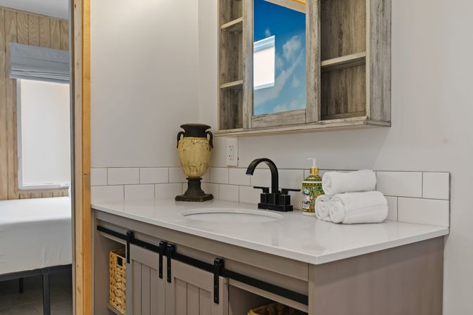 Modern farmhouse bathroom vanity with white quartz countertop, black faucet, subway tile backsplash, weathered wood medicine cabinet reflecting blue sky, rolled white towels, decorative soap dispenser and urn, sliding barn-style cabinet doors and woven baskets, glimpse into adjacent bedroom.