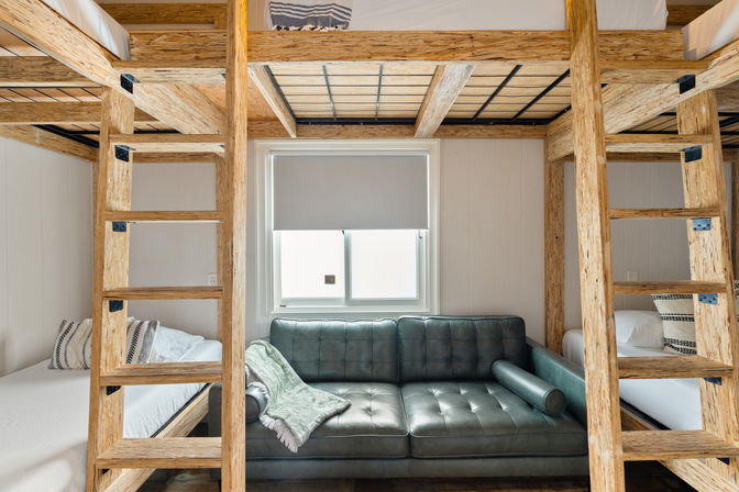 Sunlit modern-rustic bunk room with natural wood loft beds and ladders framing a dark gray tufted leather sofa under a window, flanked by twin lower beds with pillows and a soft green throw.