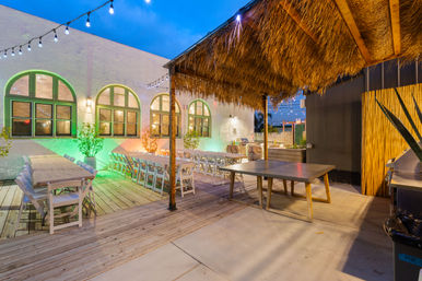Cozy evening outdoor dining patio with a wooden deck, long banquet tables and white folding chairs beneath string lights and a thatched tiki roof beside arched windows.