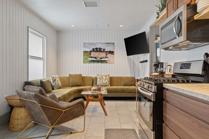 Cozy open-plan living room and kitchen with green velvet L-shaped sofa, gray accent chair with gold frame, wooden coffee table with snacks, wall-mounted TV, stainless steel gas stove and microwave, white shiplap walls and framed vintage truck photo.