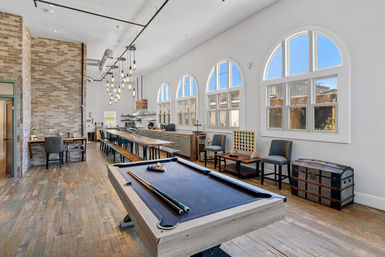 Bright loft-style communal lounge with navy pool table in foreground, long wooden communal table and benches, industrial pendant lights, exposed brick wall, arched windows, kitchenette, seating area and vintage trunk on hardwood floors.