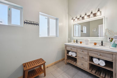 Bright spa-style bathroom with double-sink wooden vanity and illuminated mirror, open shelves with rolled white towels, wall-mounted 'TOWELS' hook, small wooden bench, and frosted windows.