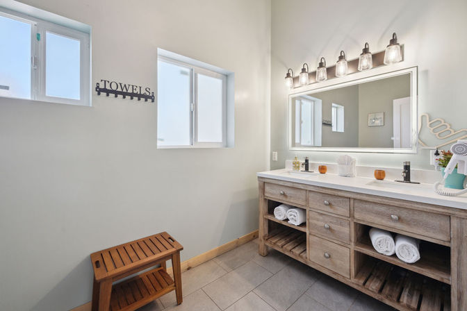 Bright spa-style bathroom with double-sink wooden vanity and illuminated mirror, open shelves with rolled white towels, wall-mounted 'TOWELS' hook, small wooden bench, and frosted windows.