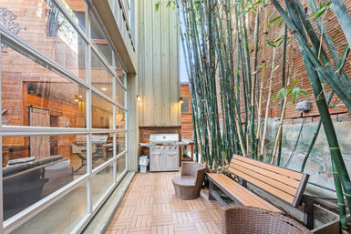 Cozy urban courtyard with tall bamboo along a brick wall, wooden bench and wicker chairs, stainless steel grill, and large glass garage-style wall opening to a modern interior.