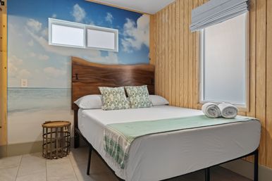 Cozy beach-themed bedroom with ocean wall mural, live-edge wooden headboard, queen bed with white linens and mint throw, patterned palm pillows, rolled towels at the foot, wood-paneled walls and frosted windows.