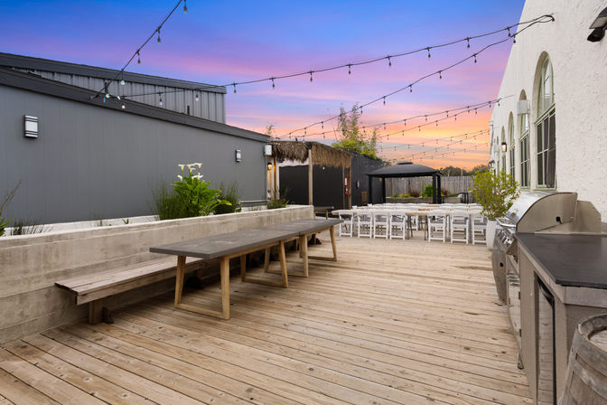 Outdoor wooden deck patio with long communal table, white folding chairs, stainless grill and twinkling string lights at sunset